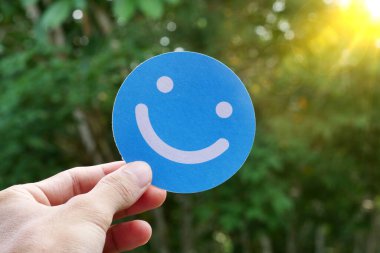 Bir insanın eli açık havada mavi bir gülen yüz ikonu tutarken güneşli, yeşil bir arka plana karşı