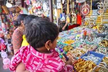 Nanded, MH, Hindistan, 21 Aralık 2025: Kırmızı gömlekli küçük bir çocuk, renkli bilezikler ve Sih dinsel hediyelik eşya satan bir tezgahta merakla taşınıyor.