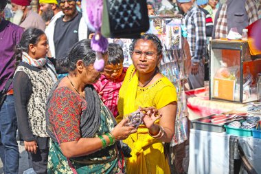 Nanded, MH, Hindistan, 21 Aralık 2025: Güneşli bir günde Sih hacılar geçerken renkli geleneksel elbise alışverişi yapan Hintli kadınların yer aldığı işlek bir sokak pazarı.