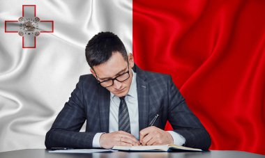 Ceketli ve gözlüklü bir işadamı masaya oturur ve Malta bayrağının arka planına karşı sözleşme imzalar.