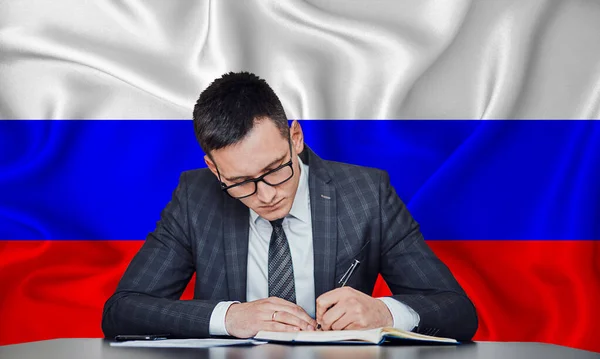 一位身穿夹克和眼镜的商人坐在桌旁 在俄罗斯国旗的背景下签署了一份合同 — 图库照片