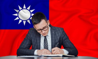 Ceketli ve gözlüklü bir işadamı masaya oturur ve bir Tayvan bayrağının arka planına karşı sözleşme imzalar.