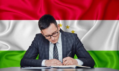 Ceketli ve gözlüklü bir işadamı masaya oturur ve Tacikistan bayrağının arka planına karşı bir sözleşme imzalar.