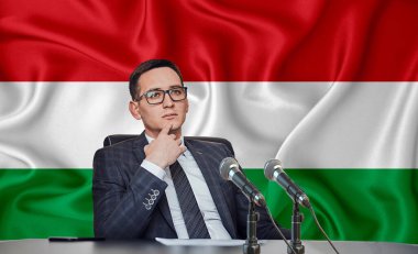 Macaristan bayrağının arka planında, uluslararası bir toplantıda veya basın toplantısı görüşmesinde gözlüklü ve ceketli genç bir adam