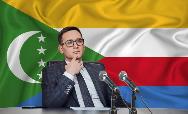 Young man in glasses and a jacket at an international meeting or press conference negotiations, on the background of the flag comores