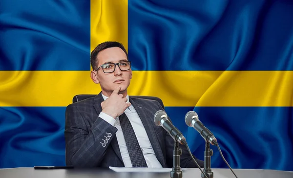 Young man in glasses and a jacket at an international meeting or press conference negotiations, on the background of the flag Sweden