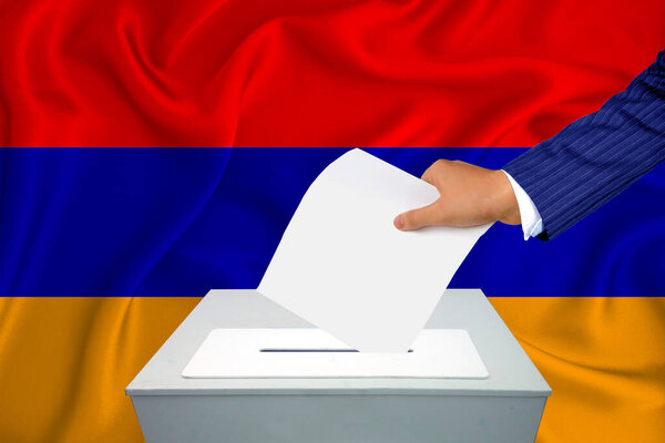 Выборы в стране - голосование у избирательного ящика. Мужская рука кладет свой голос в урну для голосования. Флаг Армении на заднем плане.