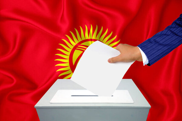 Выборы в стране - голосование у избирательного ящика. Мужская рука кладет свой голос в урну для голосования. Флаг Кыргызстана на заднем плане.