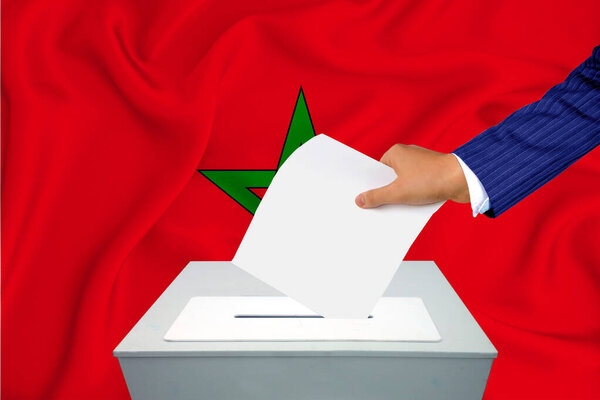 Выборы в стране - голосование у избирательного ящика. Мужская рука кладет свой голос в урну для голосования. Флаг Марокко на заднем плане.