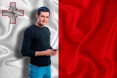 Arka planda Malta bayrağı var. Genç adam gülümsüyor ve elinde bir akıllı telefon tutuyor. Tasarım çözümleri kavramı.