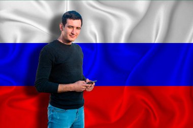 Arka planda Rusya bayrağı var. Genç adam gülümsüyor ve elinde bir akıllı telefon tutuyor. Tasarım çözümleri kavramı.