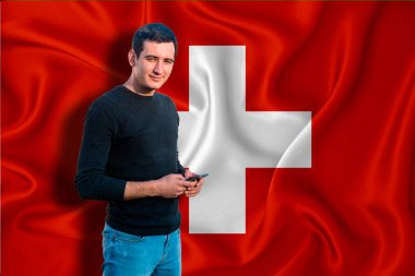 Arka planda İsviçre bayrağı var. Genç adam gülümsüyor ve elinde bir akıllı telefon tutuyor. Tasarım çözümleri kavramı.