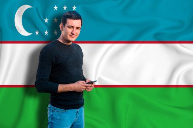 Özbekistan bayrağı dokunun arka planında. Genç adam gülümsüyor ve elinde bir akıllı telefon tutuyor. Tasarım çözümleri kavramı.