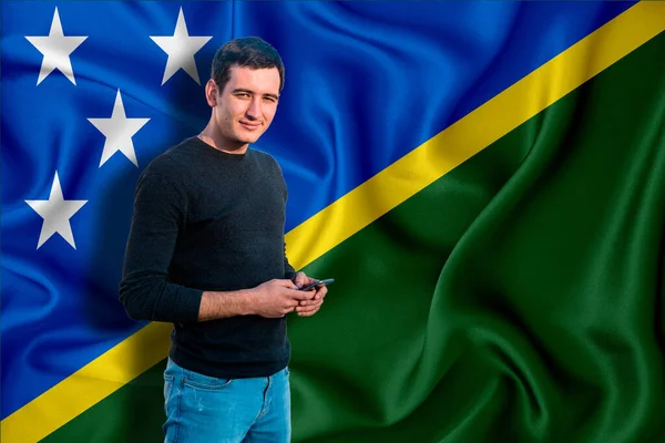 Solomon Islands flag on the background of the texture. The young man smiles and holds a smartphone in his hand. The concept of design solutions.