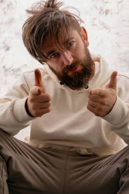 Gündelik kapüşonlu genç sakallı adam arka plandaki kameraya güvenle bakıyor, pozitiflik ve dostça tavırlar sergiliyor..