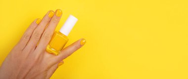 Sarı tırnak cilası tutan kadın eli. Canlı sarı arka planda izole edilmiş. Güzellik, moda ve kendine özen temaları için ideal..