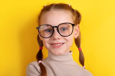 Gözlüklü ve örgülü neşeli kızıl saçlı kız parlak sarı bir arka plana gülümsüyor, bej bir kazak giyiyor. Okula dönüş veya eğitim temaları için mükemmel.