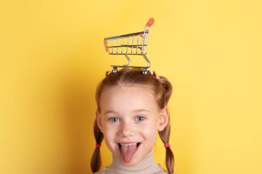 Saçları örgülü gülümseyen bir kız kafasında küçük bir alışveriş arabasını parlak sarı bir arka plan, neşeli ve eğlenceli bir konseptle dengeliyor..