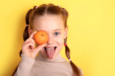 Gülümseyen genç bir kız gözüne turuncu bir mandalina tutuyor, dilini uzatıyor, parlak sarı bir arka plana karşı..