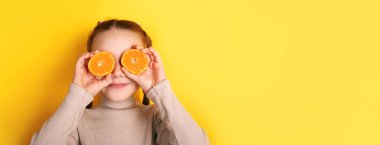 Gülümseyen kız gözlerinin üzerinde turuncu yarımlar tutuyor, parlak sarı arka plana karşı, neşeli ve neşeli bir ruh hali yaratıyor sağlık ve yaşam tarzı kavramları için mükemmel..