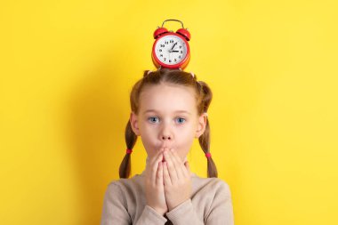 Saçları örgülü ve kafasında kırmızı alarmlı bir saat olan genç bir kız, parlak sarı bir arka plana karşı şaşkınlığını ifade ediyor..