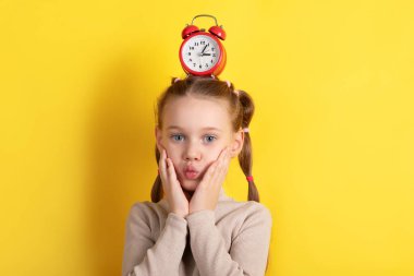 Saçları örgülü genç bir kız kafasında kırmızı alarm saatini dengeliyor, parlak sarı arka planda şaşırmış bir yüz yapıyor..