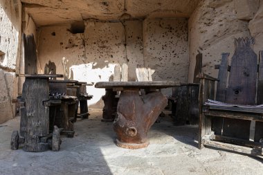 Taştan bir mağarada el yapımı ahşap masa ve sandalyeler. Sıcak güneş ışığında doğal dokular ve geleneksel tasarım sergileniyor..
