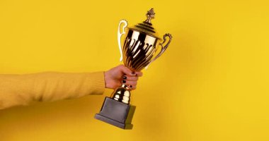 Parlak sarı bir arka planda altın bir spor kupasını tutan bir kadının yakın çekimi. Zafer, başarı, başarı, rekabet ve ödül kavramları için ideal temiz, minimal 4K video.