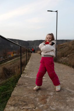 Beyaz ceketli ve pembe pantolonlu neşeli genç kız taştan bir yolda dikiliyor arka planda kış manzarası.