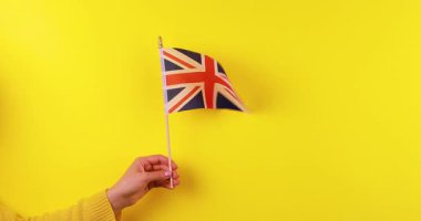 İngiliz bayrağını (Union Jack) parlak sarı arka planda tutan bir elin 4K stok görüntüsü. Temiz ve canlı kompozisyon bu videoyu vatansever içerik için ideal kılıyor.,