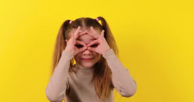 4K stok görüntülerinde beyaz bir kız parmaklarıyla eğlenceli dürbünler yapıyor ve onları parlak sarı bir arkaplanda inceliyor..