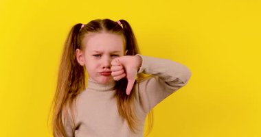 Kafkasyalı bir kızın 4K görüntüsü önce hoşlanmadığını gösteriyor, sonra gülümsüyor ve sarı arka plana başparmak işareti veriyor, ruh hali değişikliğini ve olumlu duyguyu simgeliyor..