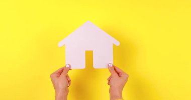4K görüntülerinde ev, mülk, konut veya güvenlik kavramlarını temsil eden sarı arka planda bir ev sembolünü tutan bir el görülüyor..