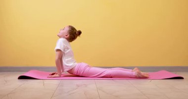 Küçük beyaz bir kız minderde yoga yapıyor, yukarı bakan köpek performansı sergiliyor ve parlak turuncu bir duvara karşı aşağı bakan köpek pozları veriyor. Bu canlı 4K videosu çocukların sağlığını ön plana çıkarıyor,