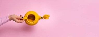 El ele tutuşan parlak sarı çaydanlık pastel pembe arka planda tek sarı lale, minimal ve modern kompozisyon..