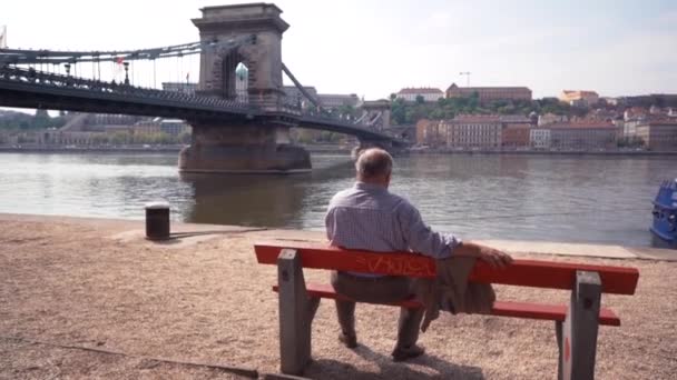 Starý muž sedí sám na lavičce a při pohledu na řece — Stock video