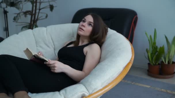 Mujer joven en casa sentada en una silla moderna, leyendo libro y sueños — Vídeo de stock