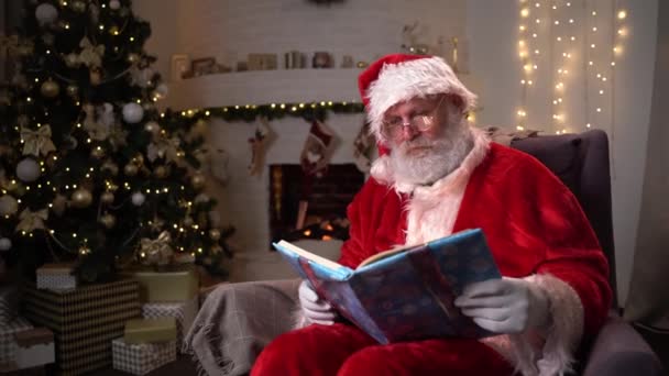 Забавный Санта оговорка сидит рядом с камином и рождественская елка чтение волшебной книги с блестящими страницами - дух Рождества, магия, мечта концепции 4k кадров — стоковое видео