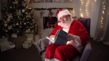 Şöminenin yanında oturan komik Noel Baba maddesi ve parlayan sayfaları olan sihirli bir kitap açan Noel Baba, sihir, rüya konsepti 4k video.