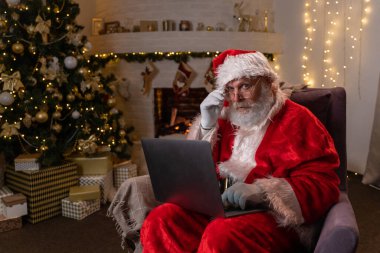 Modern Noel Baba. Neşeli Noel Baba dizüstü bilgisayarda çalışıyor ve gülümsüyor. Arka planda şöminesi ve Noel Ağacı olan sandalyesinde oturuyor..