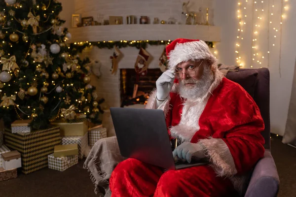 Papá Noel moderno. Alegre Santa Claus trabajando en el ordenador portátil y sonriendo mientras está sentado en su silla con chimenea y árbol de Navidad en el fondo. Imagen De Stock