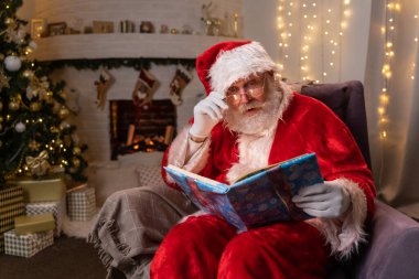 Komik Noel Baba maddesi şöminenin yanında duruyor ve noel ağacı üzerinde parlayan sayfalar olan sihirli bir kitap okuyor - Noel ruhu, sihir, rüya konsepti