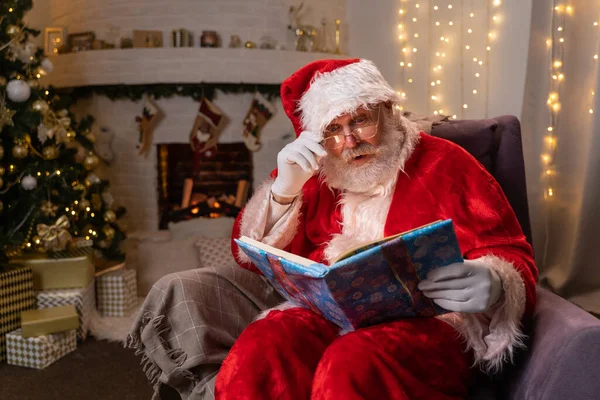 Śmieszna klauzula Mikołaja siedzi obok kominka i choinki czytając magiczną książkę ze świecącymi kartkami - duch Bożego Narodzenia, magia, koncepcja marzeń — Zdjęcie stockowe