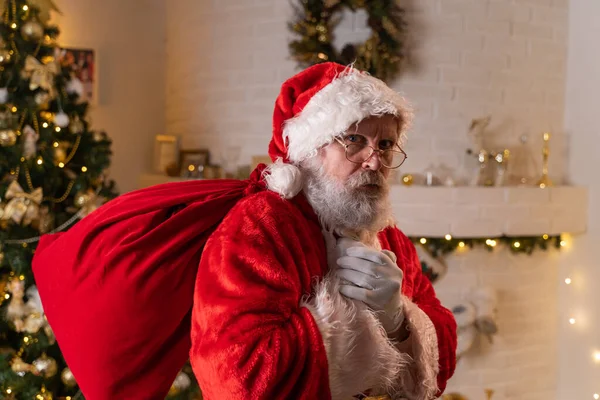 Tradycyjny Święty Mikołaj trzyma torbę z prezentami w domu w pobliżu choinki i kominka. Duch Świąt, magia, koncepcja marzeń — Zdjęcie stockowe