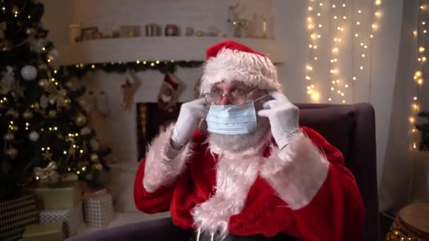 Lucunya Santa Claus mengenakan topeng medis pelindung sambil duduk di kursi dekat perapian dan pohon Natal. Covid 19, perlindungan terhadap coronavirus. — Stok Video