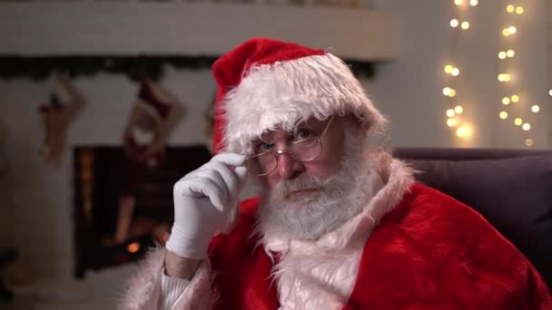 Retrato de Papai Noel engraçado em óculos, sentado em sua cadeira de balanço e coloca o dedo na boca Shhh perto da árvore de Natal. Natal, feriados e celebração conceito com resolução 4K — Vídeo de Stock