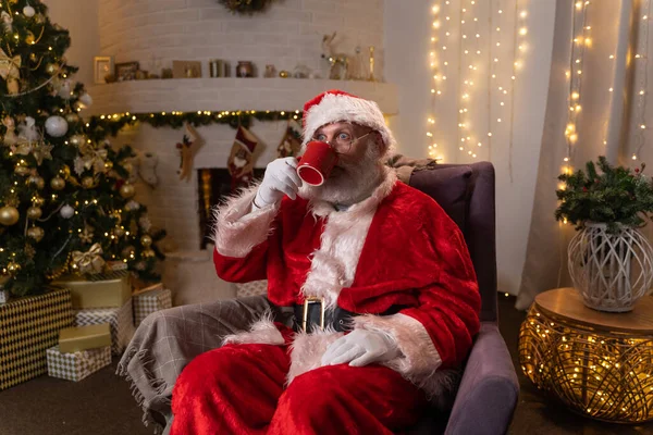 Tradycyjny Święty Mikołaj siedzi na krześle i pije ciepły napój z kubka w pobliżu choinki i kominka w pokoju urządzonym na Boże Narodzenie. Koncepcja Bożego Narodzenia i Nowego Roku. — Zdjęcie stockowe