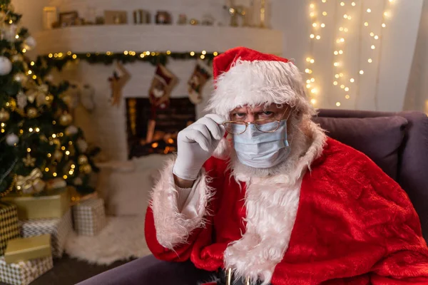 Święty Mikołaj w ochronnej masce medycznej, siedząc w fotelu przy kominku i choince. Covid 19, ochrona przed koronawirusem. — Zdjęcie stockowe