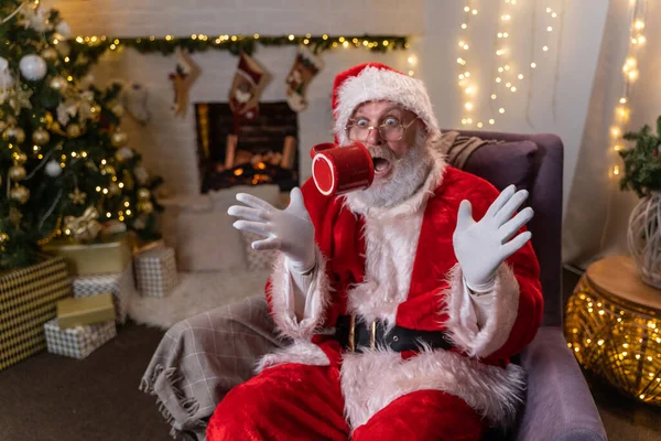 Śmieszny Mikołaj siedzi na krześle i żarty z kubkiem w pobliżu choinki i kominkiem w pokoju urządzone na Boże Narodzenie. Koncepcja Bożego Narodzenia i Nowego Roku. — Zdjęcie stockowe