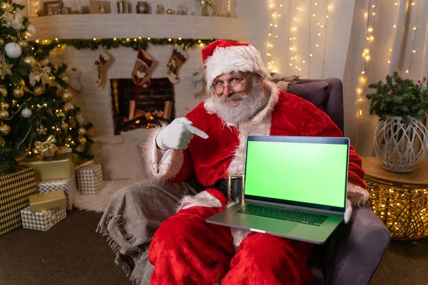 Santa Claus memakai kacamata duduk di ayamnya dekat pohon natal. memegang laptop dengan layar hijau dan menarik perhatian. Semangat Natal, liburan dan perayaan konsep rekaman 4k Stok Foto Bebas Royalti